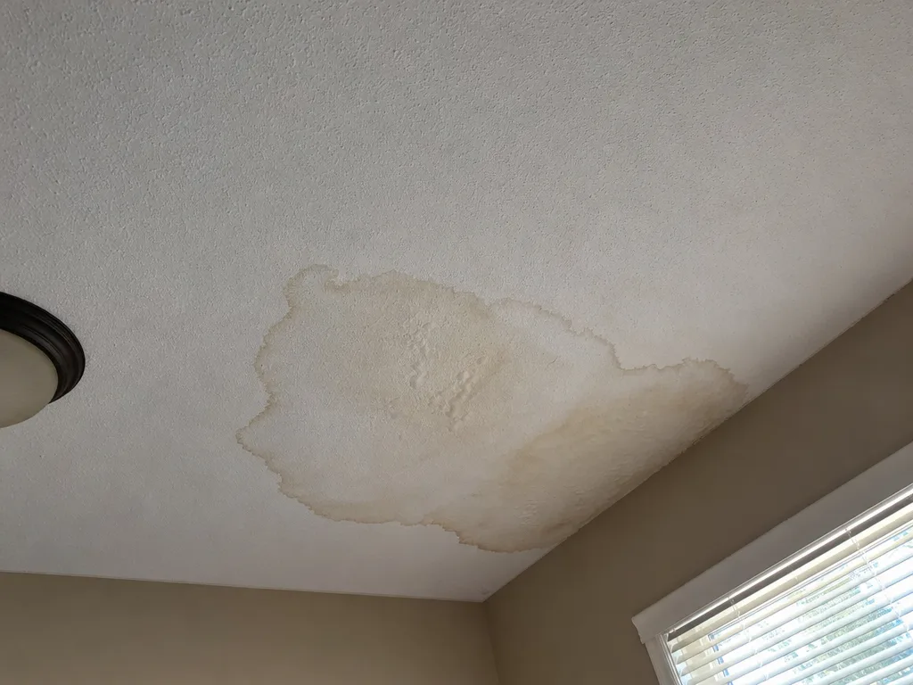 Water stain spreading across a bedroom ceiling near the window, a sign of a roof leak (illustration)