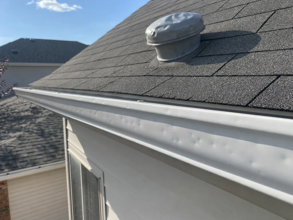 Hail dents along a white eavestrough and on a roof vent above it (illustration)