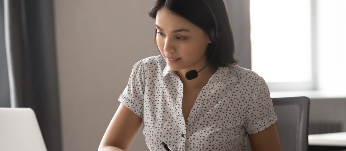 Asian businesswoman wear headset watching webinar make call write notes